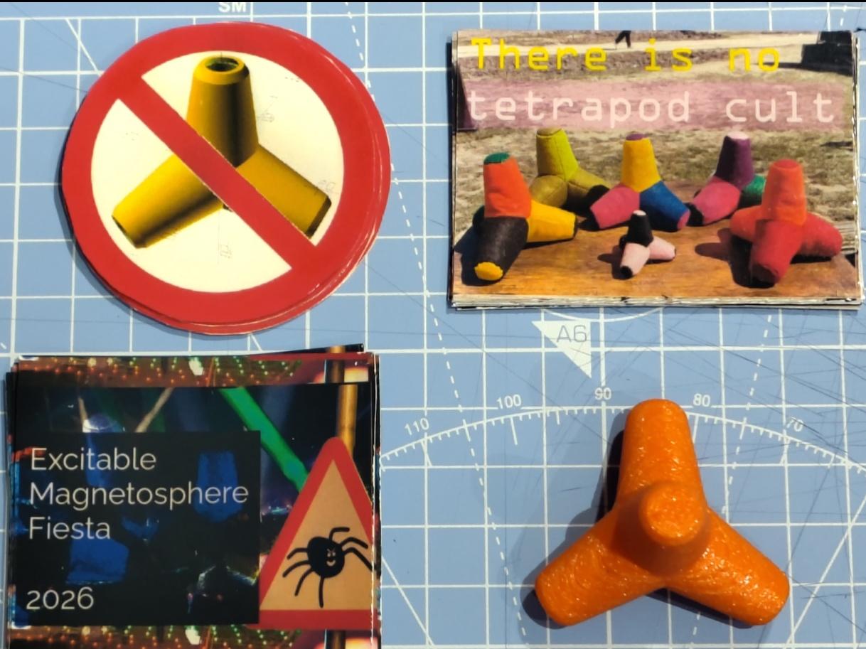 Photo showing several stickers, and something else, on a blue craft mat. One socket has a picture of several DIY plush tetrapods on a table in an outdoor setting with the title "there is no tetrapod cult". There is also a "no tetrapods" road sign style sticker showing an OpenSCAD tetrapod model behind a red circle with a line across it. Also a sticker advertising some event calling itself Excitable Magnetosphere Fiesta 2026, which appears to include copious lasers and (if you look really closely) giant tetrapods, with a warning sign featuring a cartoon spider. Finally, a small squishy emotional support tetrapod that has been 3D printed in flexible filament.