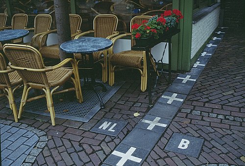 Alt4You: Picture showing restaurant tables on the Dutch side of the border, marked by tiles with “+” signs and an NL / B on either side. Photo CC BY-SA 3.0 by User:Jérôme