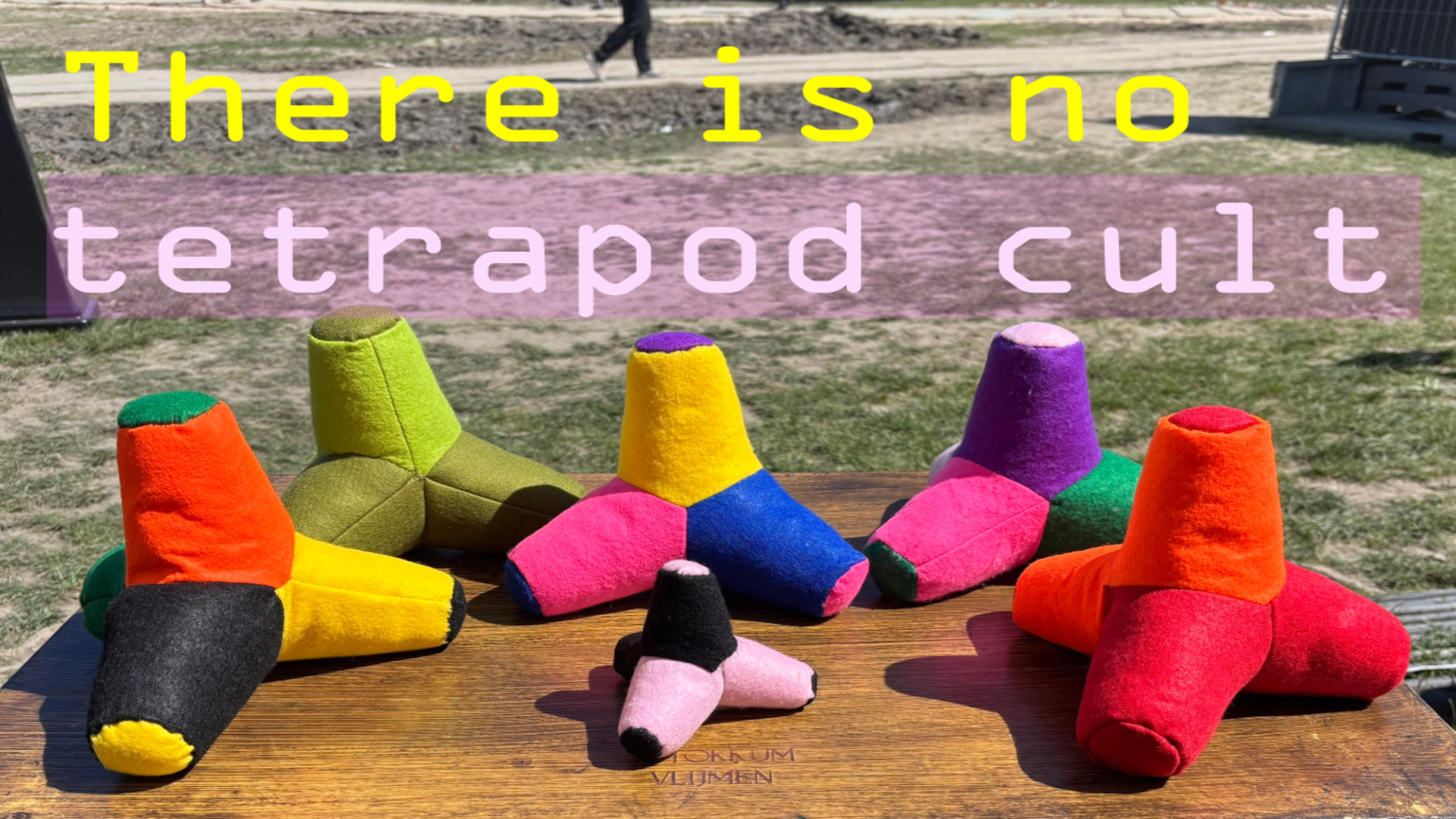 Photo showing six brightly coloured DIY plush tetrapods on a wooden table in a field somewhere in the Low Countries. Above the adorable friend-shaped items are the words ’there is no tetrapod cult’ in the OCR-A font, which was the custom at the time and is also very CYBER